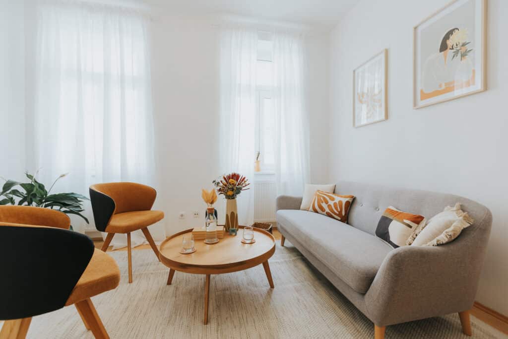 Aufnahme vom Therapie-Setting in Katharina Colombos Praxis. Ein breites, gemütliches Sofa mit zwei eleganten Lehnsessel in einem hellen, lichtdurchfluteten Raum mit hohen Fenstern und weißen Vorhängen. Am runden Couchtisch stehen Wassergläser und ein farbenfrohes Blumendekor.